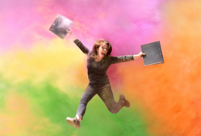 Junge Frau mit Ordner in den Händen springt freudig in die Luft vor einem bunten Hintergrund